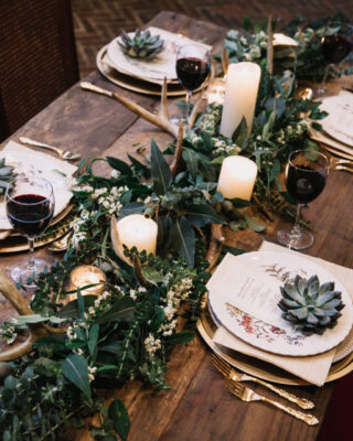 Rustic wedding table seting with vintage plates, green and white eucalyptus garland, deer antlers, gold utensils. Boho style. Table set for an event, party, date or wedding. Red wine served